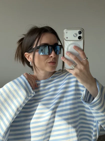 Woman taking a mirror selfie wearing blue-and-white striped top and oversized sunglasses, holding a silver phone with a clear case.