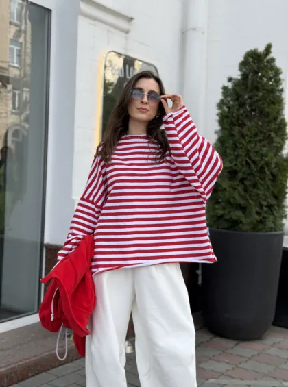 Woman in a red-and-white striped sweater and white pants, holding a red jacket, adjusting sunglasses outdoors.